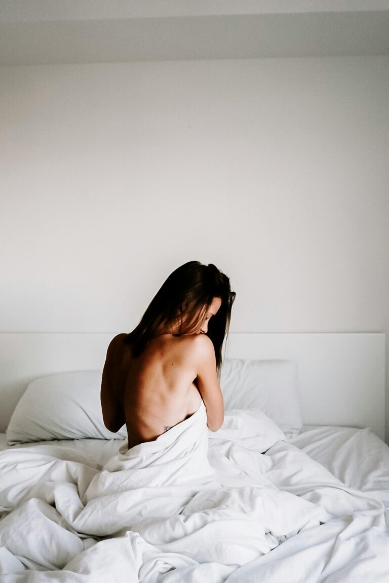 A serene, minimalist portrait of a woman sitting on a bed, enveloped in soft white linens, in a tranquil bedroom setting.